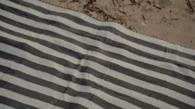 Striped towel and tote bag on beach, sand texture and pink lettering, soft morning light, minimal composition, empty sunbathing setup suggesting recent departure, relaxed summer mood