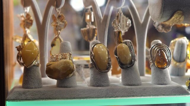 Slow close-up video of premium amber rings and pendants displayed on gray velvet stands inside a jewelry shop case, warm lit gemstone souvenirs with silver settings, retail showcase, no people