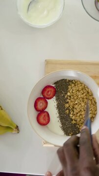 Vertical video: Entering hands, man in shorts nudging slices with fork to make yogurt bowl at home