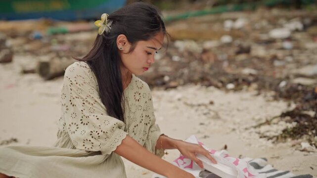 Asian woman on beach packing container into bag, moves with calm purpose, gathers items from shoreline, places container into reusable bag, beached boat in background, floral hair clip, lace dress,