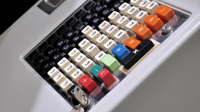 Angled close-up of an old mechanical cash register keypad with numbered black and cream keys, colorful function buttons and plastic cover, vintage retail payment machine detail, no people