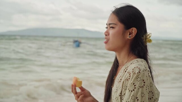 Female enjoys fresh seaside bounty. Woman delicately tastes fresh produce by tranquil shoreline and boat. Lady silently savors juicy fruit while relaxing by seaside with waves and boat nearby