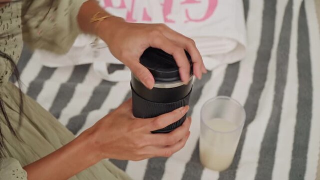 Asian woman on beach preparing iced drink on striped towel. Plastic tote and glass of milk beside her, hands opening tumbler lid and adding ice cubes. Closeup captures steady hands and simple ritual,