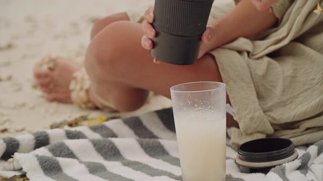 Beach towel coffee cup, caucasian woman pours from reusable mug beside frothy glass on sand, sunlit toes and bracelet visible, actions suggest digital nomad or travel writer taking coffee break, calm