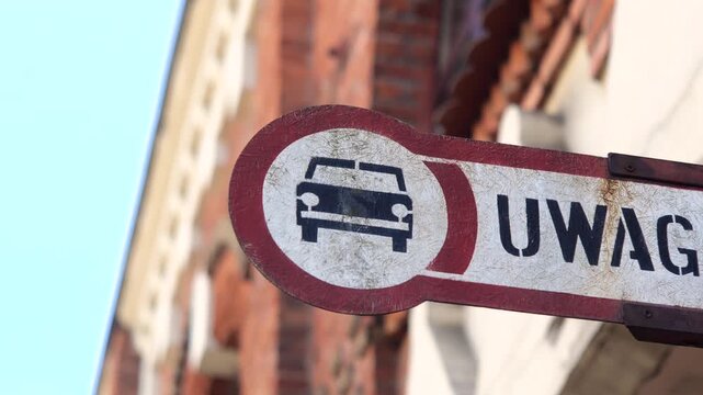 Weathered old caution sign with car symbol and Polish warning text mounted on a building facade, close-up of restricted vehicle access notice with blurred urban background, no people