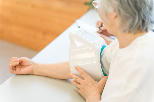 A senior woman uses a blood pressure monitor to measure her blood pressure, while a nurse and caregiver record the results.