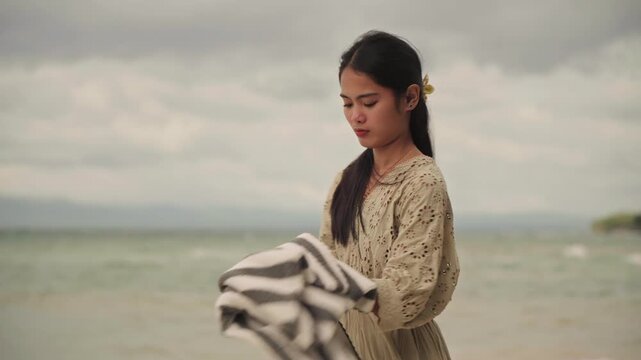 Asian woman folding striped towel beach, braided hair in loose plait, vintage eyelet dress, overcast sky with gentle waves, calm reflective mood, slow hands smoothing fabric, closeup and medium shots