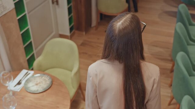 Caucasian woman entering empty cafe scanning tables and plates, green chairs and wooden flooring visible, wearing blazer and long straight hair, planning reservation and seating layout with calm