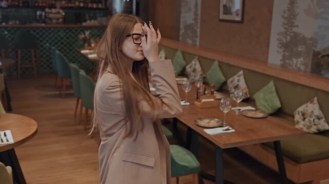 Woman in cozy dining room walks between tables scans seating arrangements, checks reservations, adjusts handbag strap, smiles toward staff, explores private venue with stage and microphone, warm wood