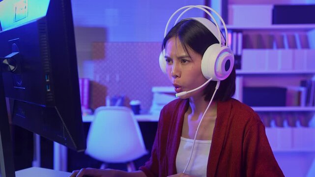 Excited young Asian girl playing online computer game with headset and microphone at home, esports and live streaming concept.