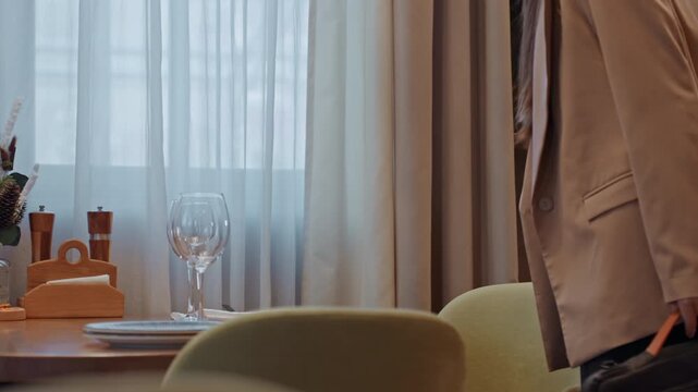 Black woman entering cozy restaurant space carrying bag and adjusting blazer, poised stride toward table with wine glasses, modern interior, soft daylight through curtains, expectation of meeting,