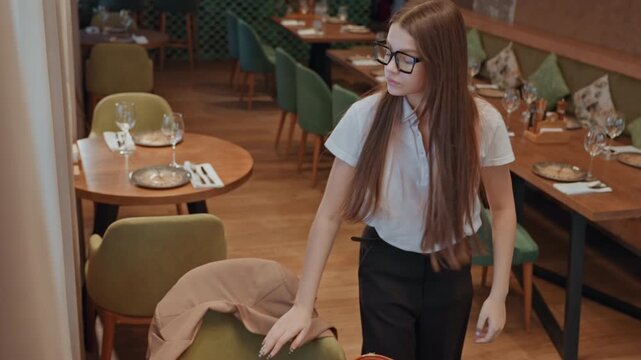 Caucasian woman waiting alone at table, empty restaurant interior with green chairs and wooden tables, glasses on face, laptop bag on table, checks phone and bag, pulls on jacket and walks away, mood