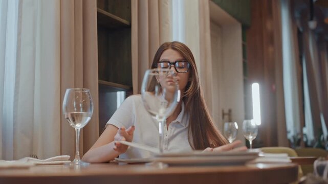 Caucasian woman sitting at restaurant table reading menu wearing glasses long hair waiting to order sipping water under warm ambient light cozy bistro interior freelance editor reviewing notes