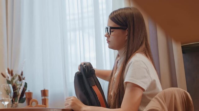 White teen girl resting on backpack at dining table, thoughtful gaze toward window, glasses, soft daylight, reaching for remote, preparing to resume laptop work, calm yet distracted mood, remote