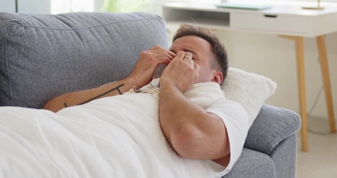 Sick man with virus cold flu and fever. At home on the sofa, scarf and blanket on, he rubs eyes and fights cough, sneeze and congestion while resting. Clear seasonal viral illness concept.