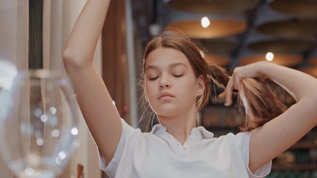 Young caucasian woman styling hair near mirror and glass, indoor cafe interior with warm lighting, hands gathering hair into ponytail, calm expression, closeup sequence showing routine grooming