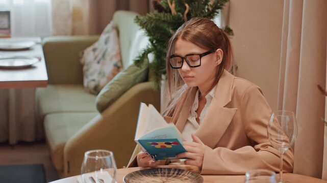 Young woman reading novel at table in cozy cafe setting, wearing blazer and glasses, soft warm light, sofa and plants in background, plate and empty wine glass on table, focused expression, slow paced
