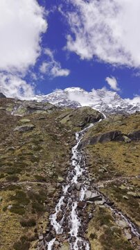 Aerial view of a mountain stream cascading down a rocky slope with snow-capped peaks under a blue sky in Alagna Valsesia, Piedmont, Italy.