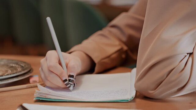 Closeup hand writing notebook, cozy cafe aesthetic, beige sleeve, patterned manicure, ceramic plate beside pages, soft bokeh background, focused study vibe, pen strokes and handwritten notes,