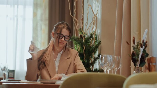 White woman at restaurant table, startup founder reading tough message, dabbing tears with napkin, covering mouth and blinking, then pausing in thought soft curtains, floral vase and glassware
