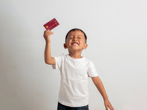 Happy young asian boy holds a red credit card near his ear while smiling broadly during this bright studio photo that captures a playful moment of childhood financial learning experiences.