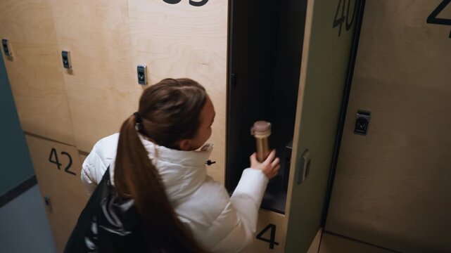 White woman opening numbered locker retrieving bag and securing coat puffer jacket, ponytail, plywood lockers, quick glance, purposeful stride through teal corridor of urban sports facility commuter