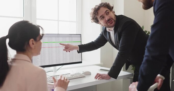 Colleagues meeting in office discuss data analytics. Manager points at a spreadsheet on the monitor to guide coworkers through metrics and tasks. Team focus on planning and strategy.