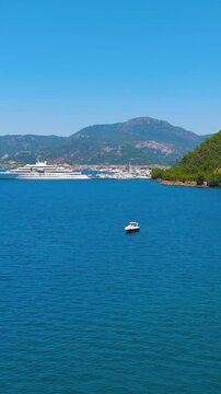 Vertical video. Marmaris, Turkey. A large white luxury motor yacht moored in a scenic Mediterranean bay, aerial drone view over deep blue water with pine-covered hills and distant mountains under bri