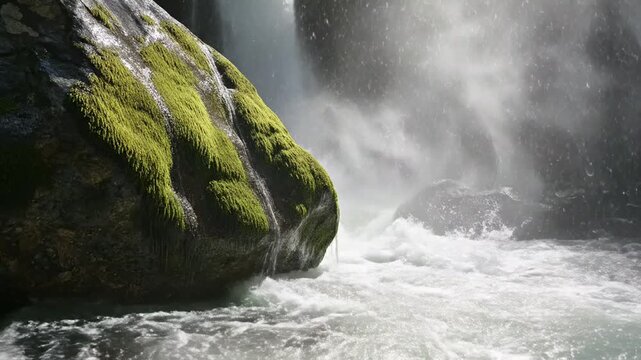 Waterfall cascades over a large mossy rock formation surrounded by mist and rushing water in a lush natural forest environment.