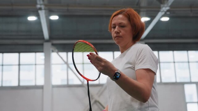 Redhaired white woman adjusting racket strings on indoor court, trainer testing tension and grip, wrist check and timing practice, natural light through windows, focused technical routine, equipment