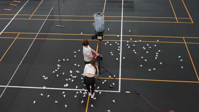 Overhead badminton court scattered with shuttlecocks, two players practicing with shuttle launcher, coach adjusting machine, one player executing smashes while partner retrieves shuttles, court lines