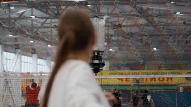 Badminton lob practice with shuttle launcher. Young player practicing lofted shots and trajectory control, automated feed sending high shuttles over net, indoor training hall with coaches observing,