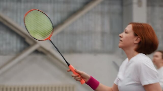 White redhead playing badminton indoors, club player driving fast forehand with wristband and bright racket, partner blurred in background, quick rally motion, determined expression and sharp reflexes