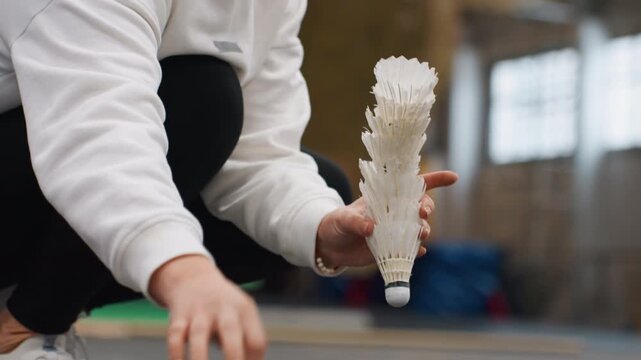 Player collecting shuttlecocks on court, hands stacking stacks of feathered shuttles and arranging pile, indoor sports hall scene with coach nearby, quick warmup and drill setup, communal practice
