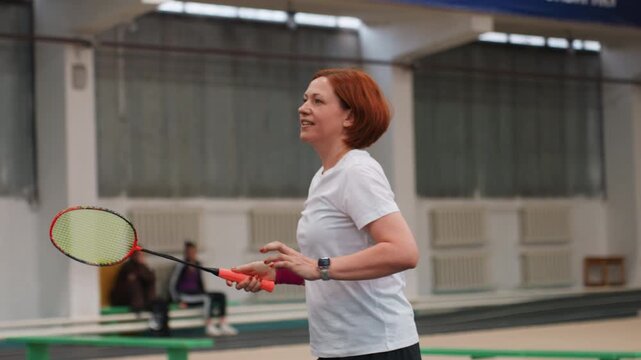 Redhead caucasian woman serving badminton indoors, focused overhead shots and wrist control, wearing pink wristband, solo drill and precision practice, coach observing sidelines, determined match