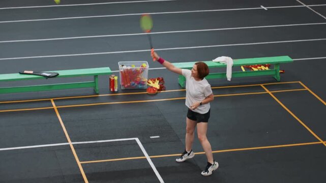 Woman playing badminton in gym, clubstyle rally with forehand and backhand returns, white tee and shorts, green benches and gear at court edge, steady footwork and playful competitive energy
