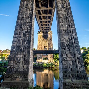 Low-angle perspective of a monumental stone bridge, featuring robust pillars and intricate girder structure. The bridge spans a calm river under a bright blue sky