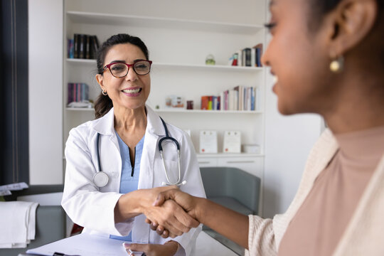 Cheerful middle aged physician shaking hands with young Black patient, giving respect, support, confidence, empathy. Healthy young African woman givin handshake to doctor, thanking for medical help