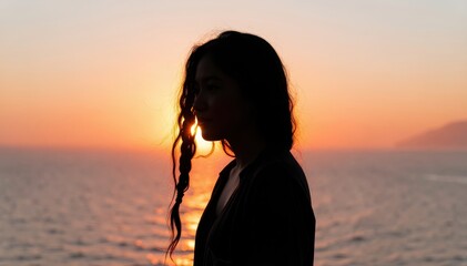 Fototapeta premium Silhouette of a Woman at Sunset: A woman's profile is silhouetted against a vibrant sunset over the ocean, her long, dark hair catching the golden light as she looks out at the serene horizon.