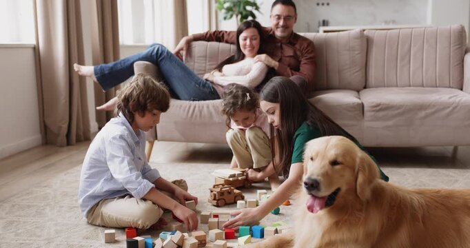 Children playing with wooden building blocks on floor while parents relaxing seated on sofa, large family spending weekend leisure in living room with golden retriever dog. Childhood games at home
