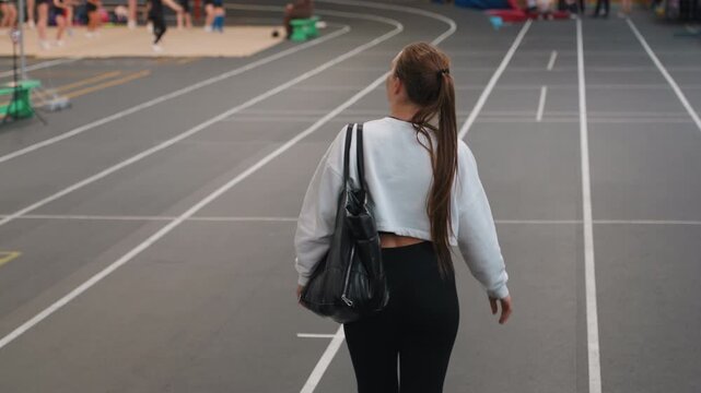 Woman walking away. Braid swings as woman departs scene. Evening lights illuminate woman ending her badminton game. Spectators fade into background as woman with braid and bag exits peacefully
