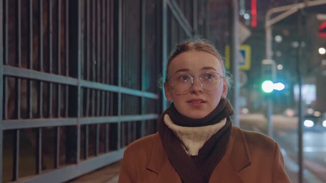 Young woman on night sidewalk recording selfie with phone, wearing brown coat and chunky scarf, round glasses reflecting neon city lights, leaning near metal railing, candid vlog and street interview