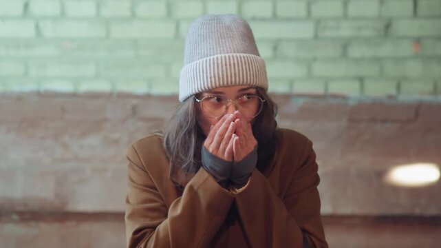 White woman sitting on bench in brick station adjusting glasses and scarf, beanie and coat layered for winter, soft vintage light and warm glow, shy smile and playful gesture suggest student waiting