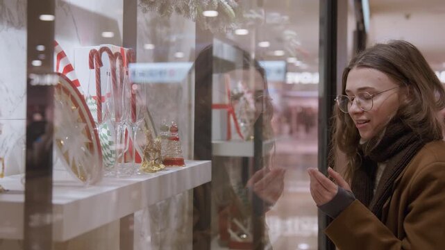 Caucasian woman admires holiday window display, gazing through glass at ornaments, candy canes, porcelain figures and gold baubles wearing coat, scarf and glasses, soft mall lights and reflective