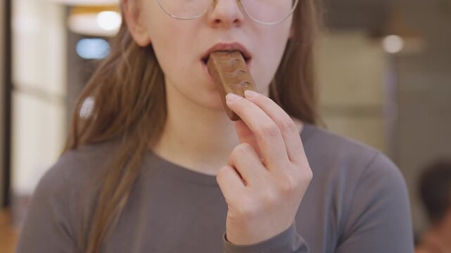 Young woman eating chocolate bar slowly, glasses and neutral sweater, warm indoor lighting, macro framing of mouth and fingers, tasting and savoring bite, candid cafe background, relaxed student mood
