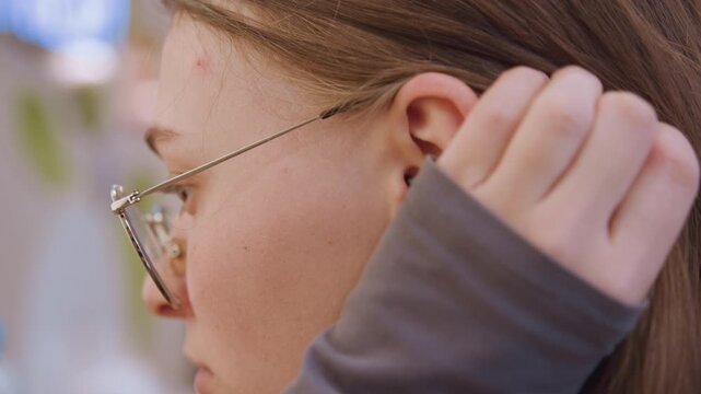 Caucasian young woman touching ear profile closeup street cafe bokeh backdrop, long brown hair tucked behind ear, metal rim glasses, subtle hand gesture, concentrated expression, rehearsal