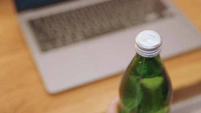 Green bottle with white plastic cap closeup of hands removing cap over wooden desk by laptop, content creator performing small ritual to open drink, tactile thread detail and quiet workspace