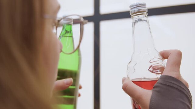 White woman inspecting green and clear bottles up close, product tester examines liquid color and label near bright window, quiet studio mood, glasses perched on face, careful hand movements, intimate