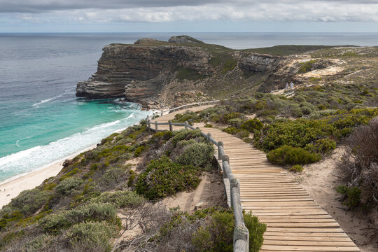Scenic view of Cape of Good Hope, South Africa, featuring a wooden boardwalk trail with handrails leading towards a dramatic rocky cliff, turquoise ocean water, and a cloudy sky over Dias Beach.