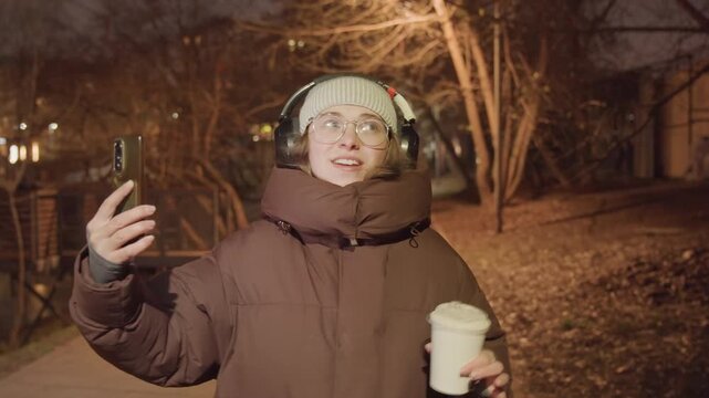 Asian influencer nighttime park selfie coffee, holding phone and cup while gesturing, wearing oversized coat, headphones and beanie, playful sip and smile, streetlight glow and leafless trees, candid